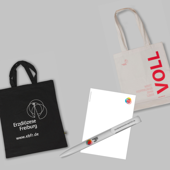 Werbeartikel der Erzdiözese Freiburg mit Stofftaschen, Notizblock und Kugelschreiber auf grauem Hintergrund arrangiert