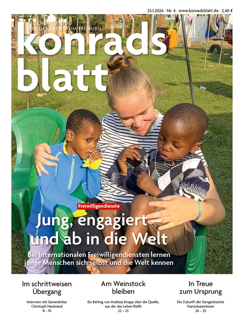 Junge Frau sitzt auf einer Wiese und umarmt zwei Kinder, im Hintergrund weitere Personen und ein Gebäude.