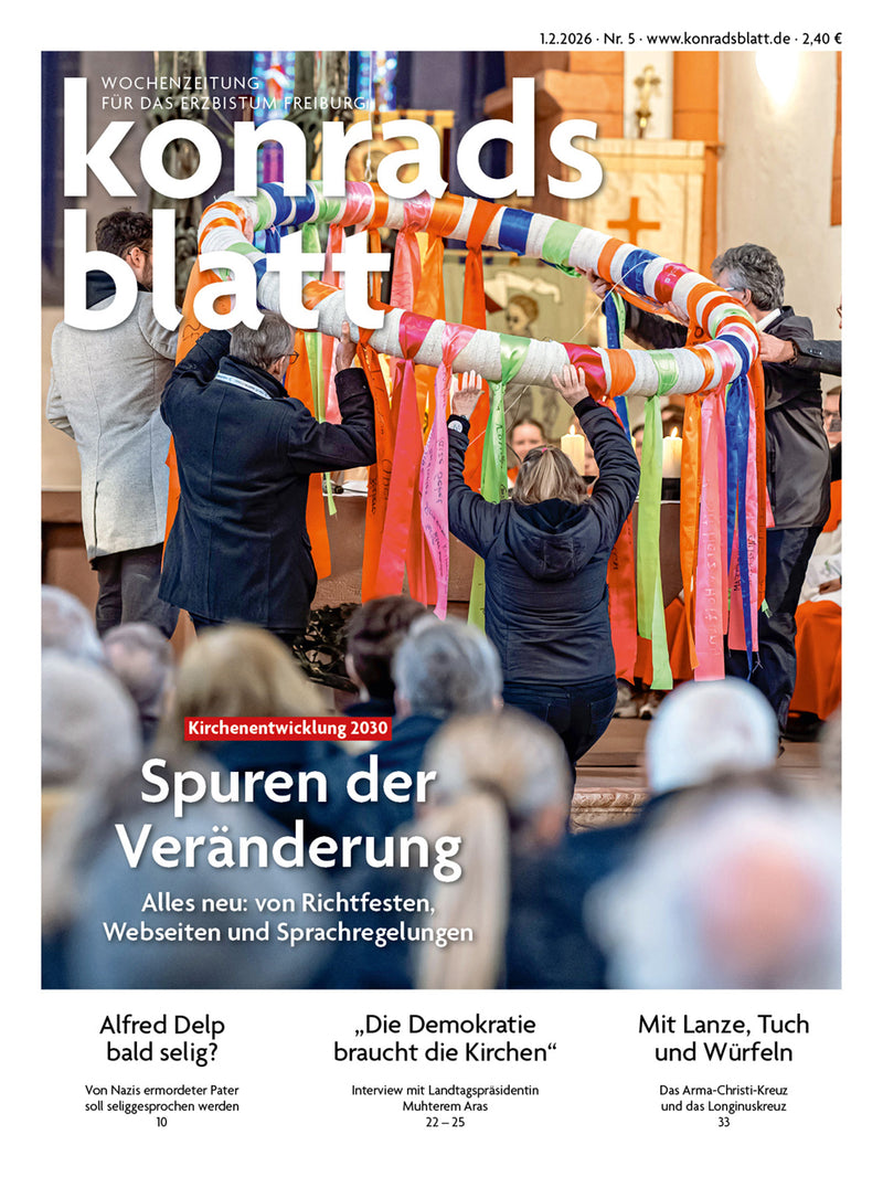 Mehrere Personen halten gemeinsam einen großen, mit bunten Bändern umwickelten Ring in einer Kirche, im Vordergrund sind verschwommene Köpfe von Zuschauern zu sehen.