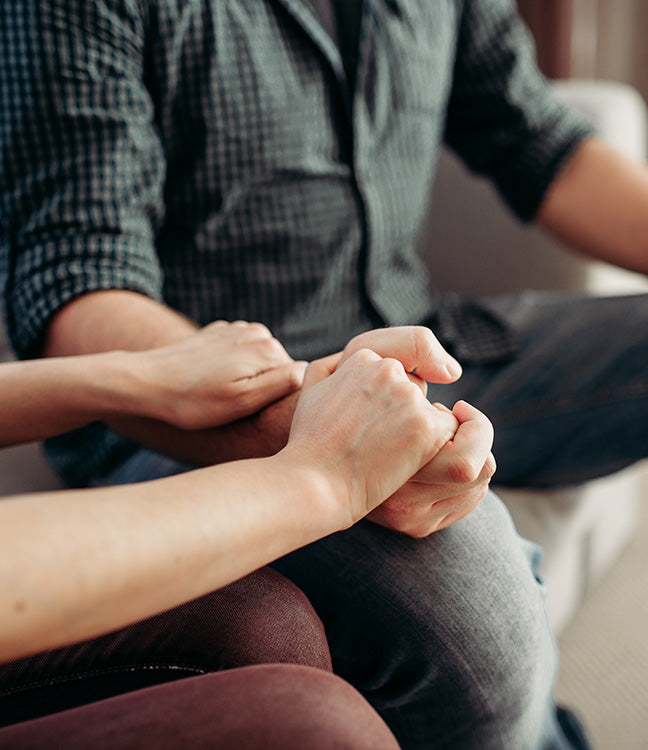 Zwei Personen sitzen nebeneinander, eine legt ihre Hand tröstend auf die Hand der anderen