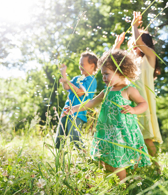 Drei Kinder spielen im Freien zwischen hohem Gras und Wildblumen an einem sonnigen Tag
