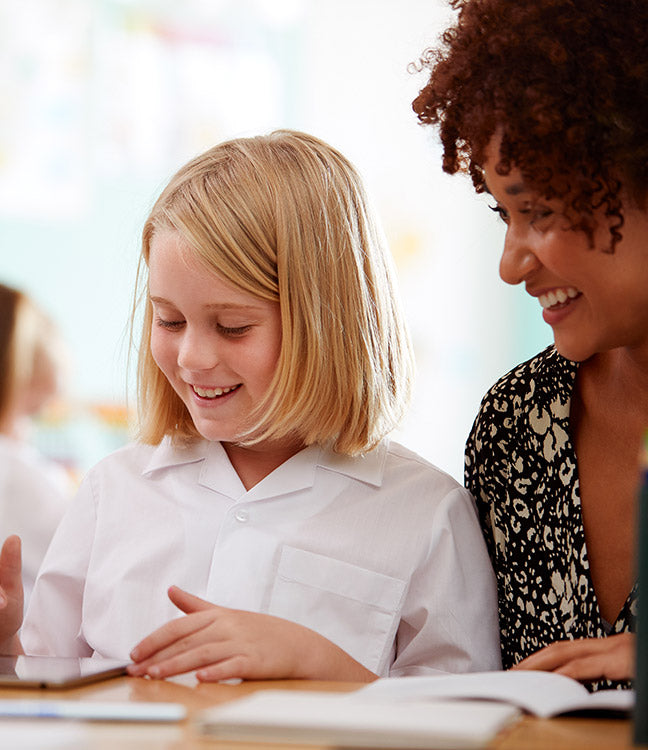 Kind mit schulterlangem blonden Haar in weißem Hemd sitzt an Tisch mit Tablet und Buch neben Person mit lockigem Haar und gemustertem Oberteil