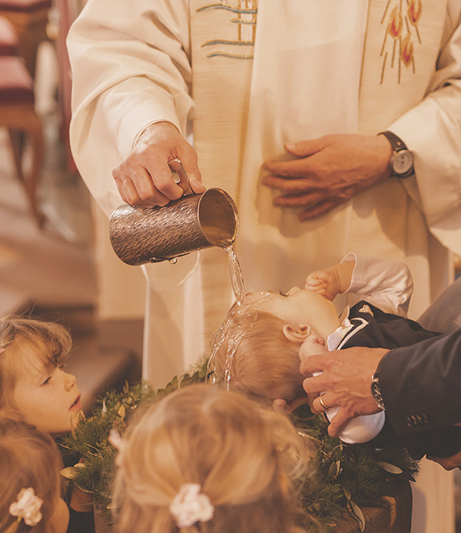 Ein Geistlicher in liturgischer Kleidung vollzieht die Taufe eines Kleinkindes, indem er Wasser aus einem Metallgefäß auf dessen Kopf gießt, während im Vordergrund zwei weitere Kinder andächtig zuschauen.